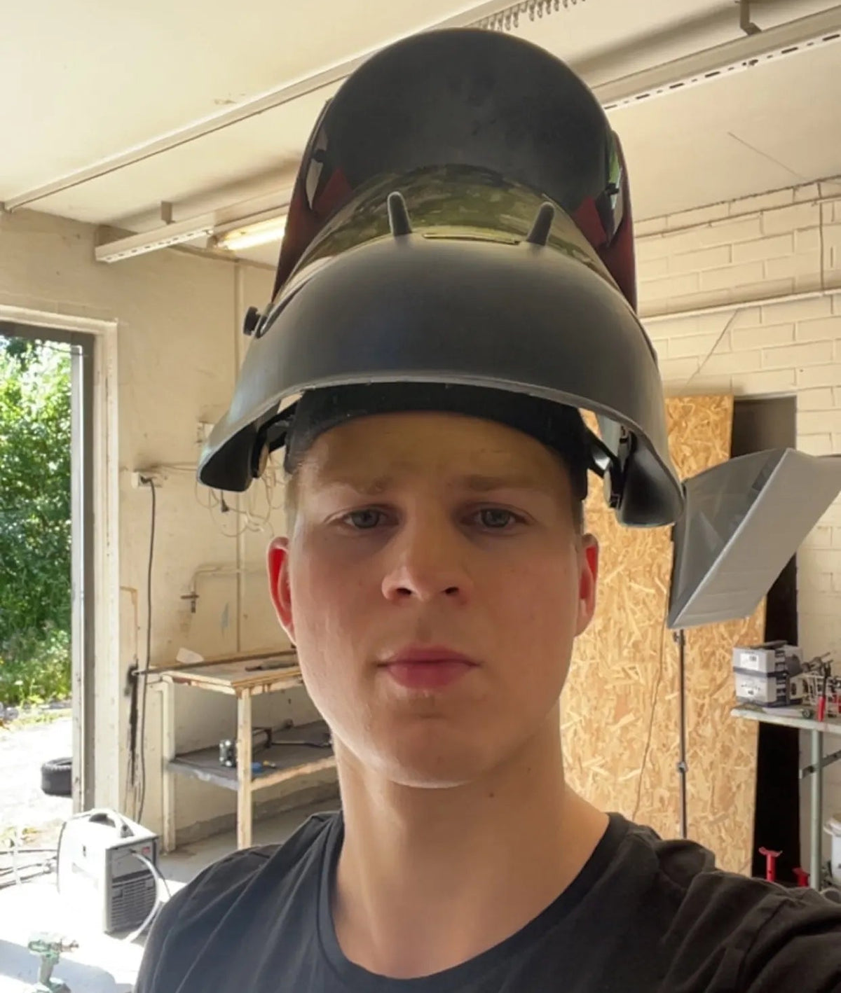 A man wearing a black welding helmet with a tinted shield in a workshop setting.